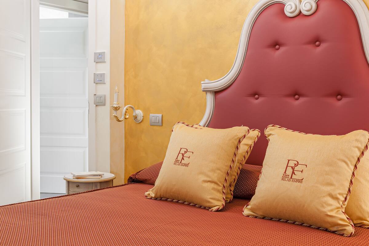 A bedroom with a red upholstered headboard, two matching pillows with a logo, a bedside table with a book, and a door leading to another room.