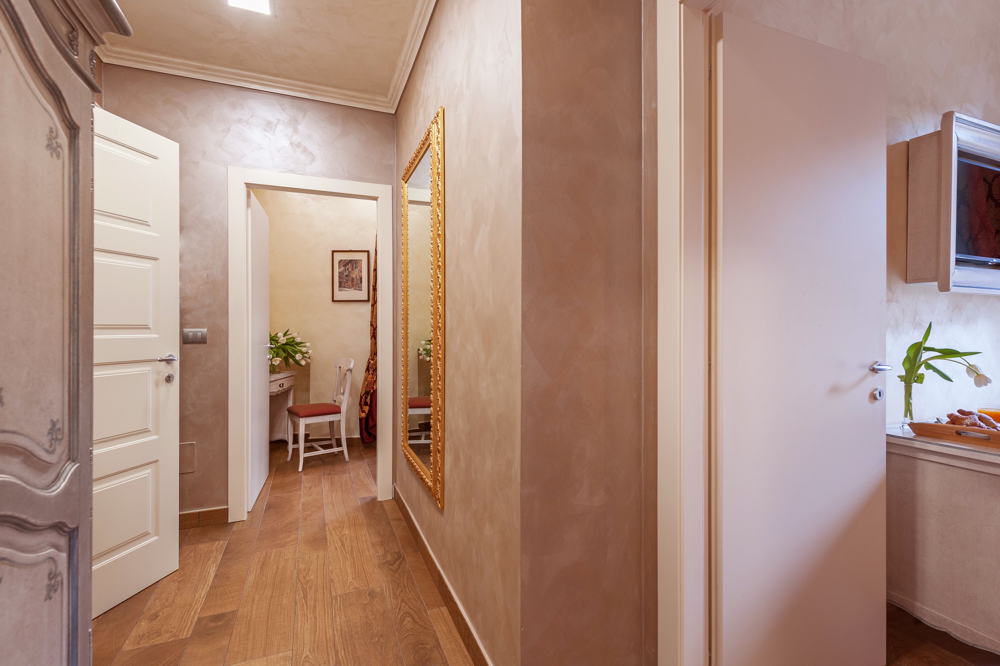 Un pasillo con piso de madera, una puerta blanca, un banco, un espejo y una pequeña mesa con una planta.