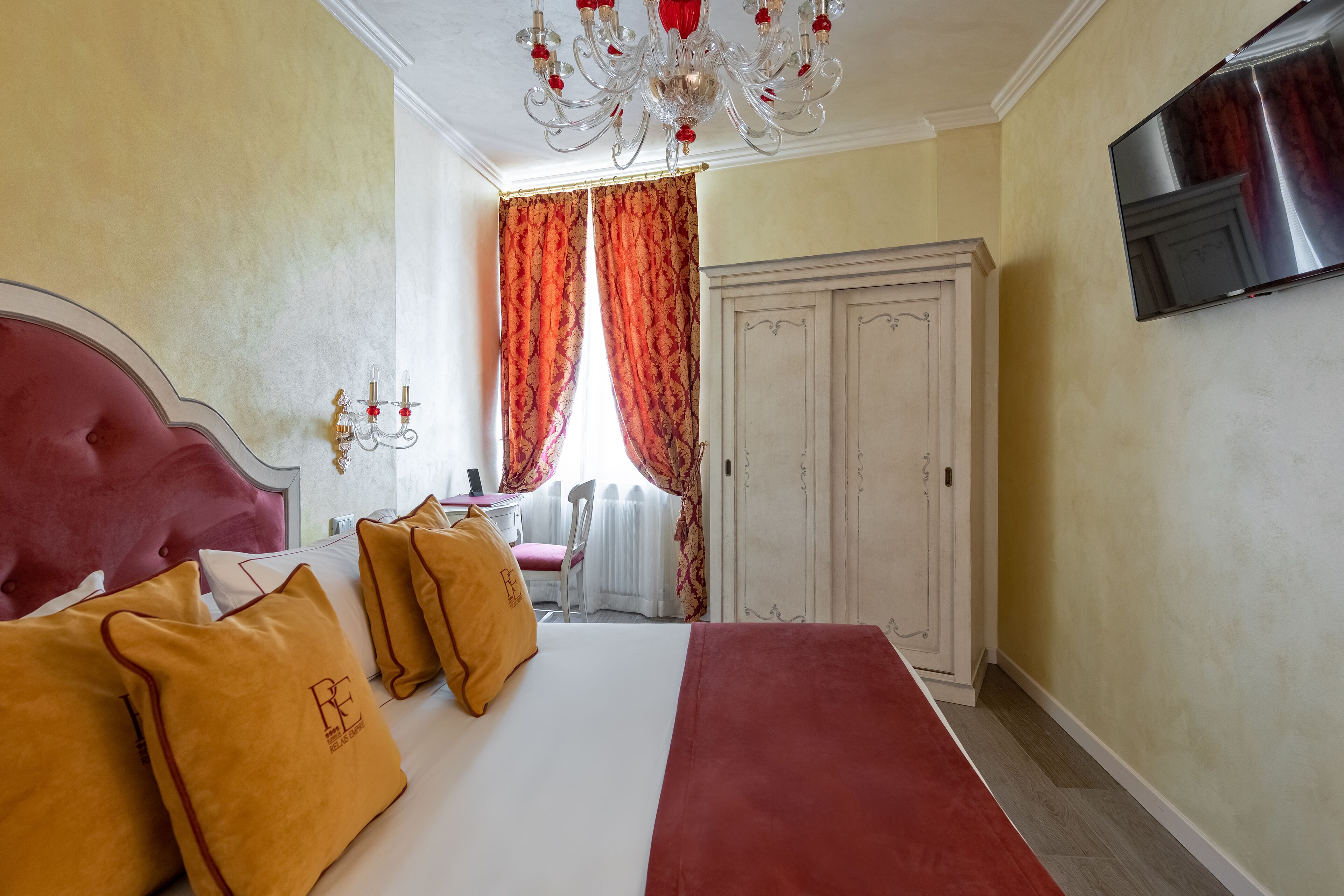 Une chambre d’hôtel avec un grand lit, des oreillers rouges et jaunes, un lustre, une armoire et un téléviseur fixé au mur.