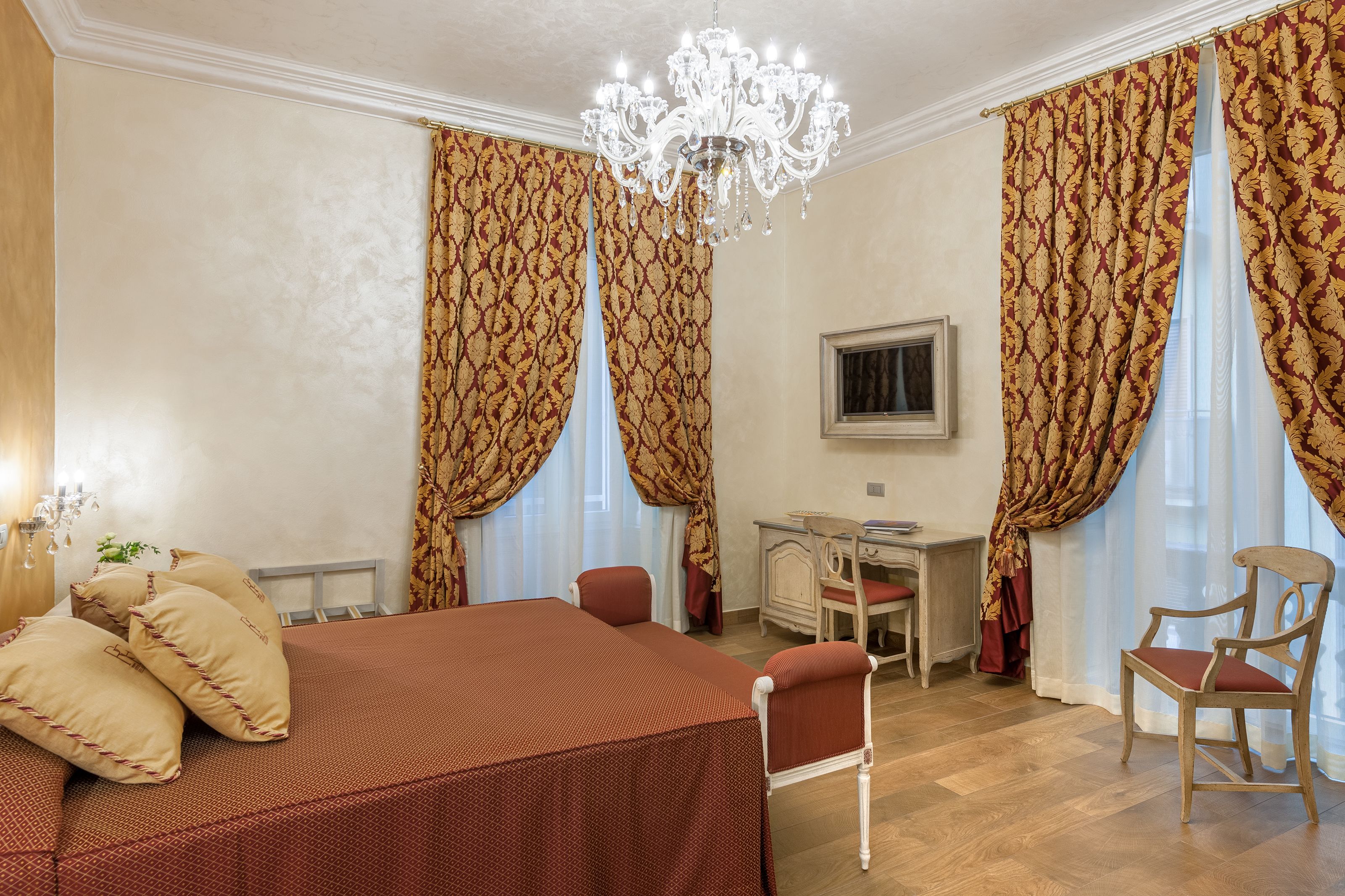 Une chambre d’hôtel avec un grand lit, un bureau, deux chaises, un lustre et des rideaux à motifs floraux.