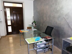 Een kamer met een glazen tafel waarop verschillende kaarten en documenten liggen, een zwarte bureaustoel, een houten deur en een wand met een grijze structuur.