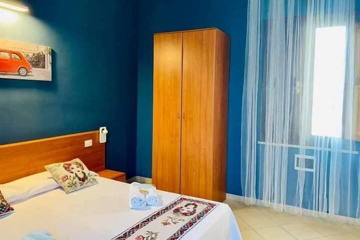 Ein Schlafzimmer mit Bett, Schrank, Nachttisch und einem Fenster mit transparenten Vorhängen.