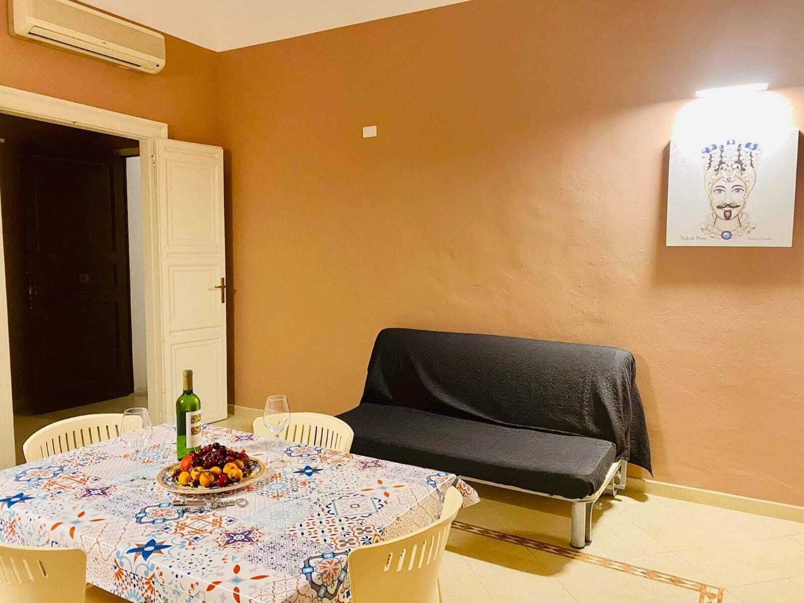 Ein Essbereich mit einem runden Tisch, der mit einer gemusterten Tischdecke bedeckt ist, einer Obstschale, einer Weinflasche und einem schwarzen Sofa.