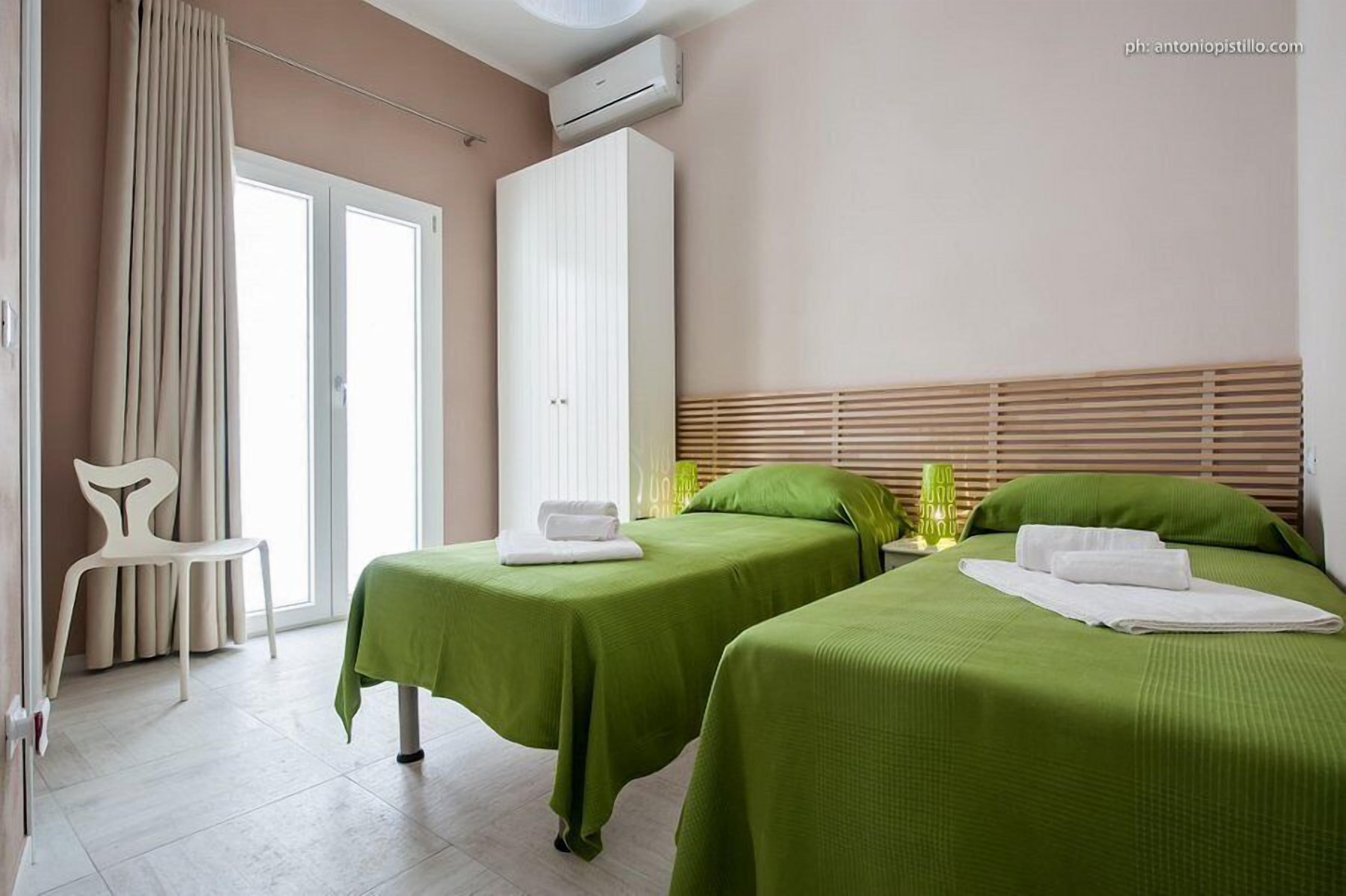 A hotel room with two beds, a chair, a wardrobe, and a window with curtains.