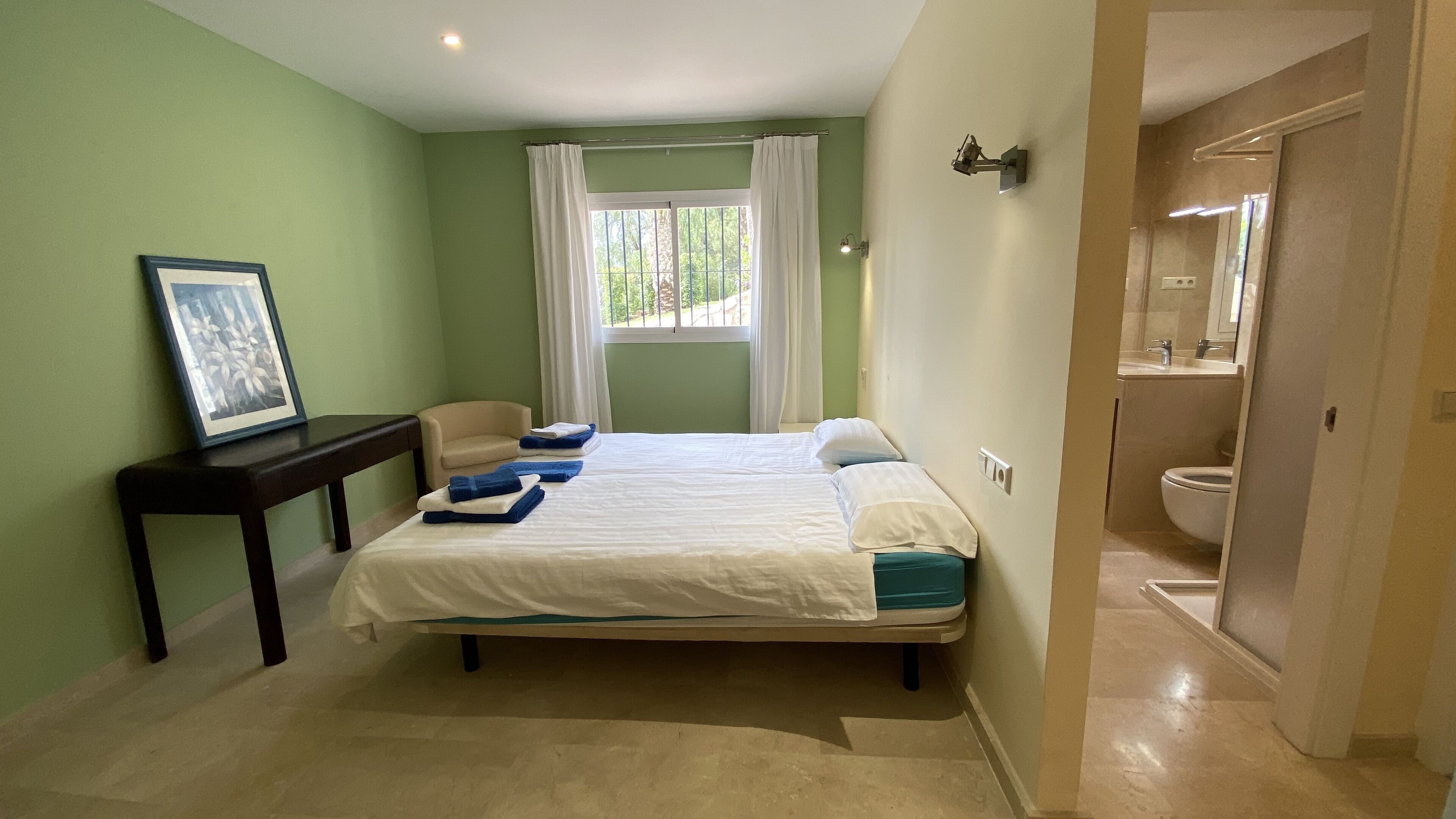 A bedroom with a bed, a bedside table, a chair, a window with curtains, and a bathroom visible through an open door.