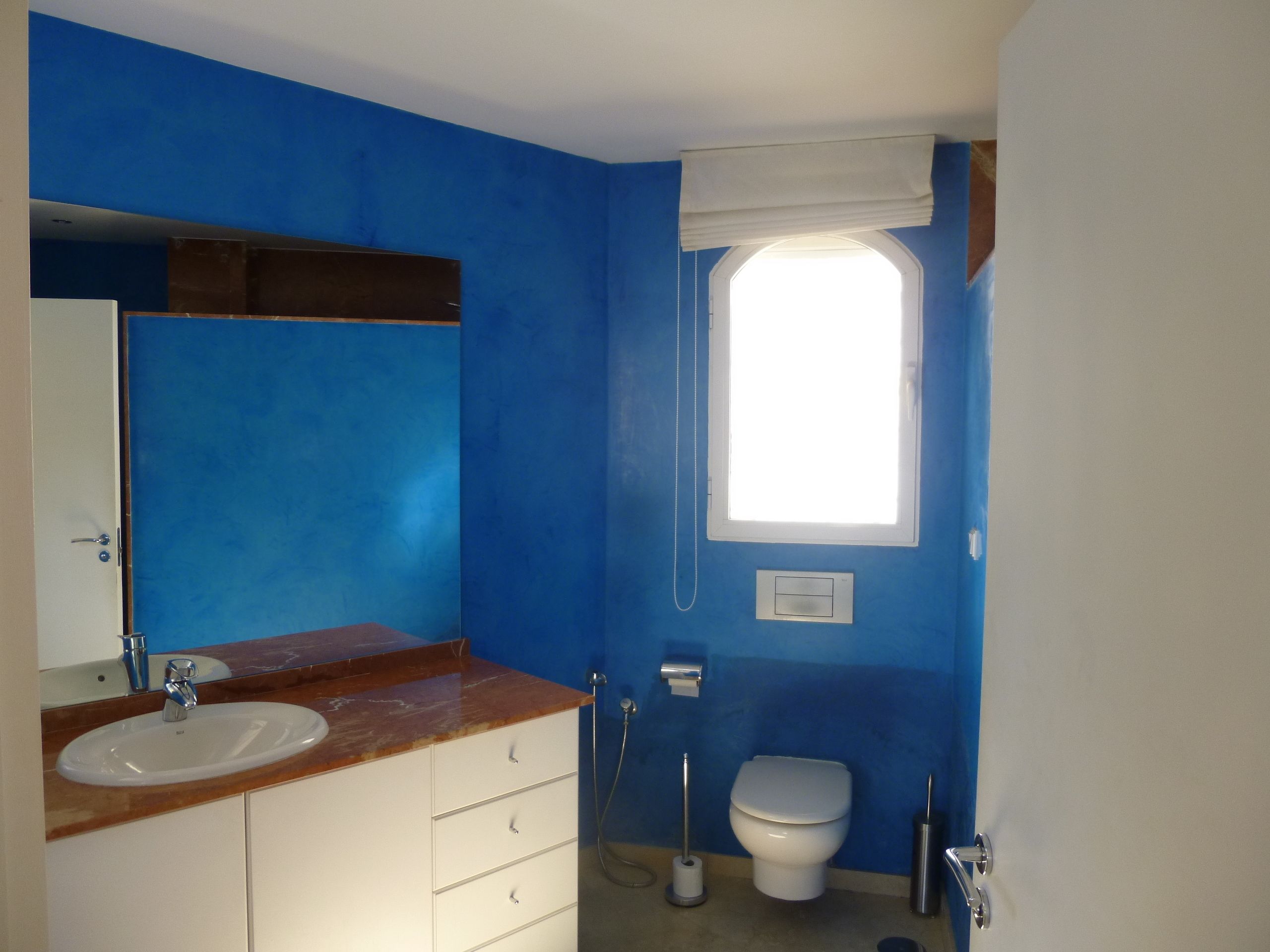 Un baño con pared azul, un lavamanos blanco, un espejo, una ventana y un inodoro.