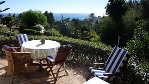 An outdoor patio with a round table and chairs, surrounded by lush greenery and a view of the sea. - La Perla de Torrenueva (Mijas)