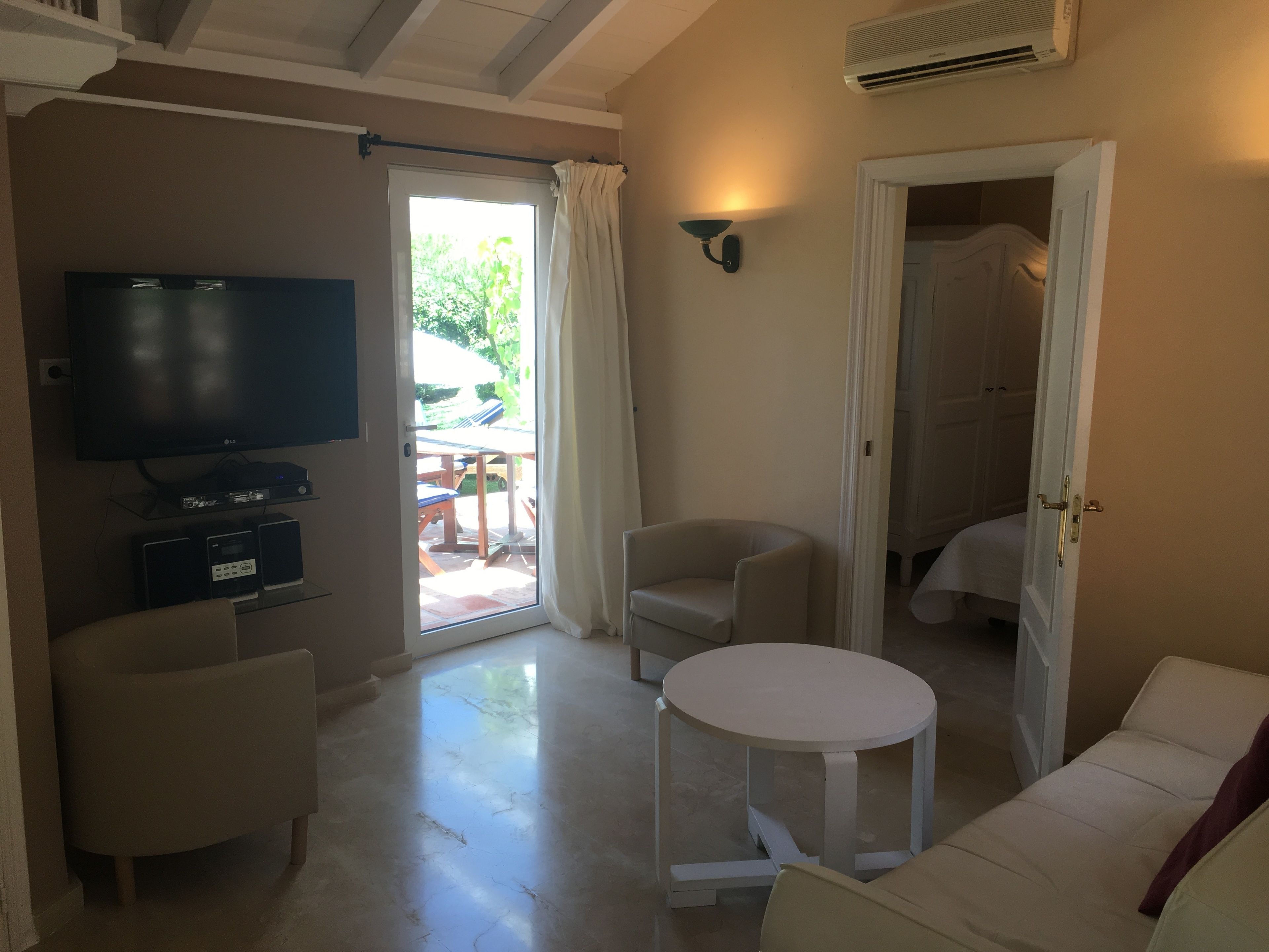 A living room with a flat-screen TV, a small table, and a sofa. There is a door leading to a balcony with outdoor furniture.