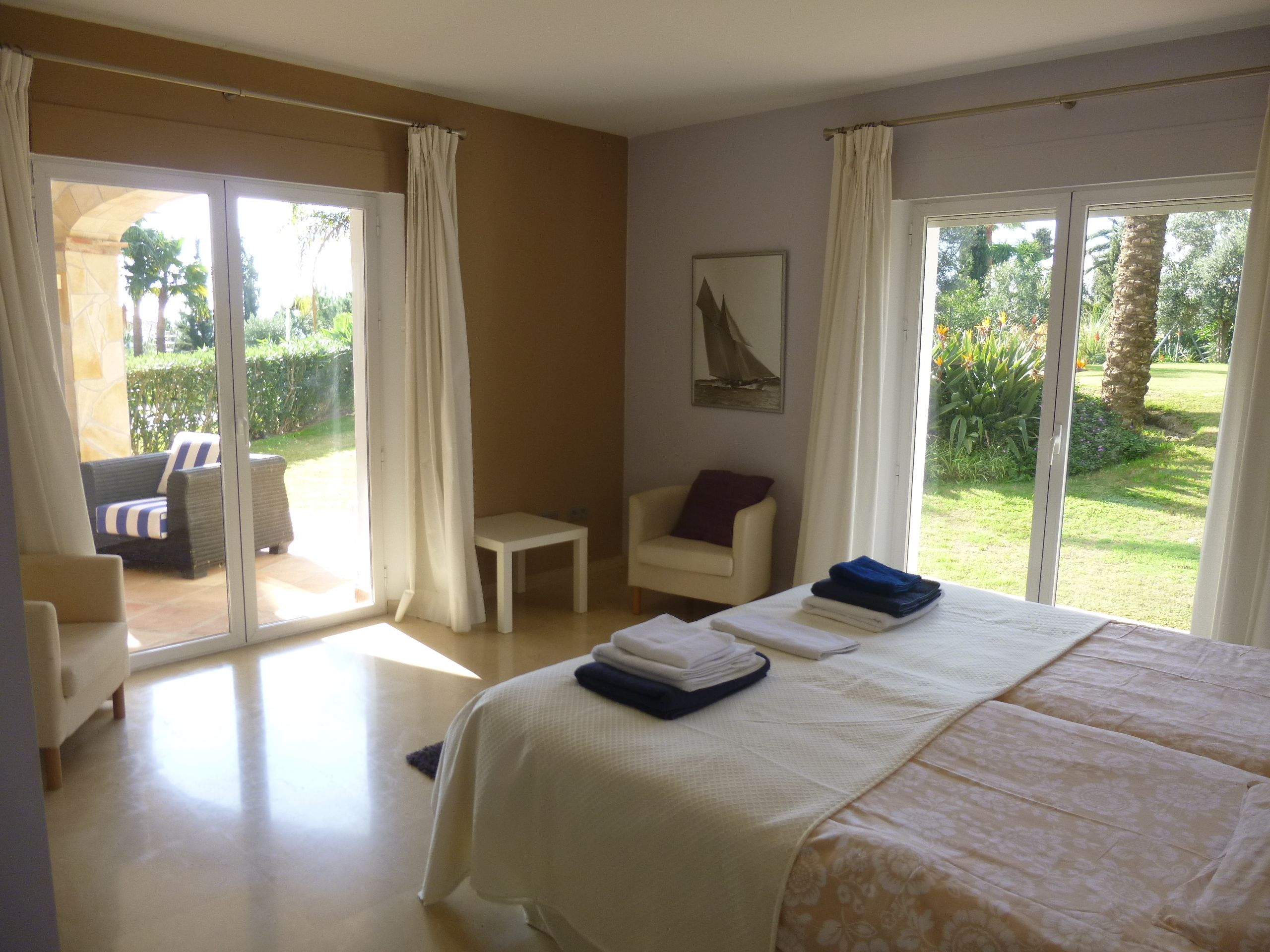 A hotel room with a bed, a chair, a small table, and a view of a garden through large windows.