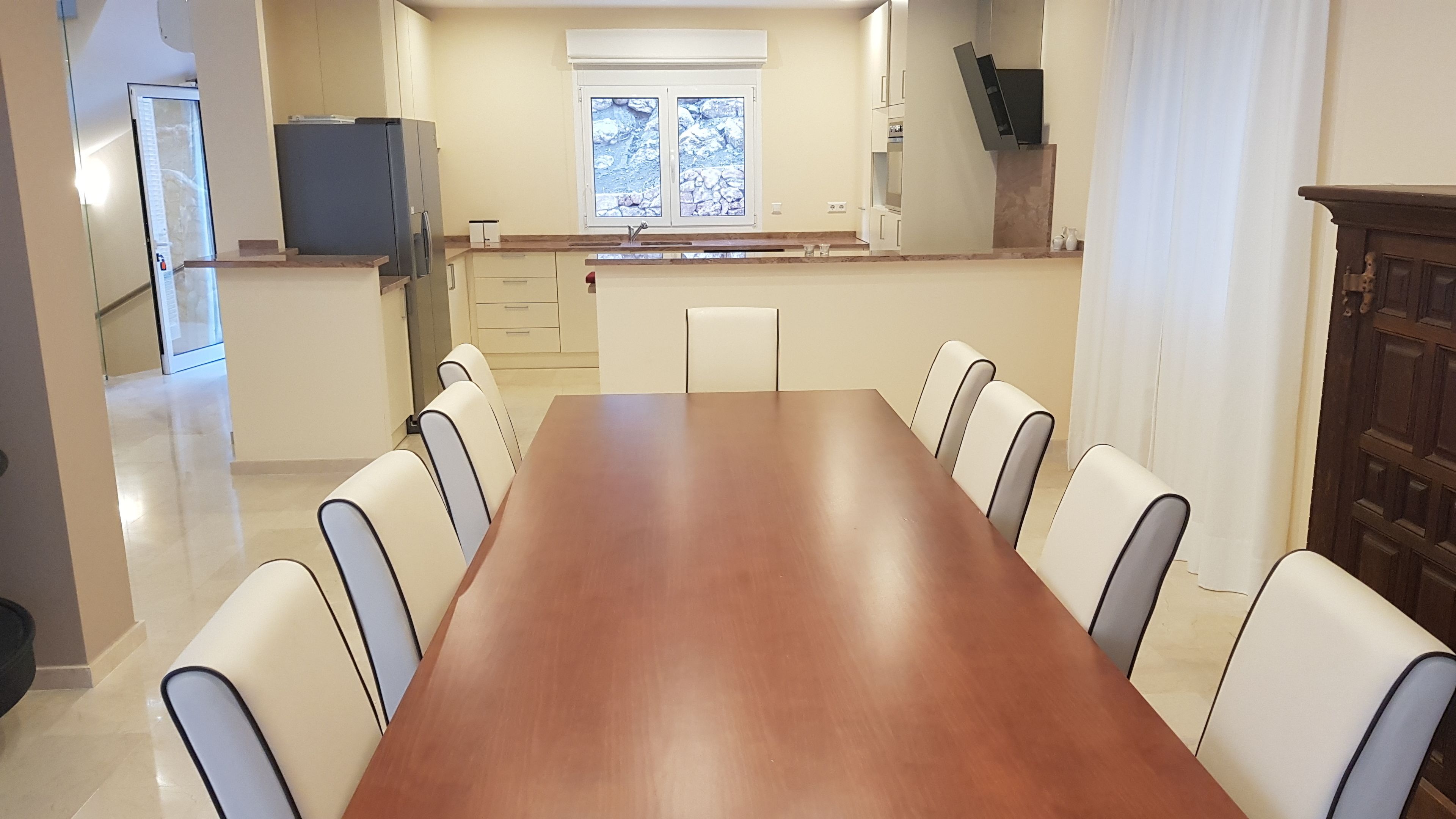 Un grand espace à manger avec une longue table en bois et des chaises blanches, une cuisine équipée d’un réfrigérateur et de placards, et une fenêtre donnant sur un paysage rocheux.