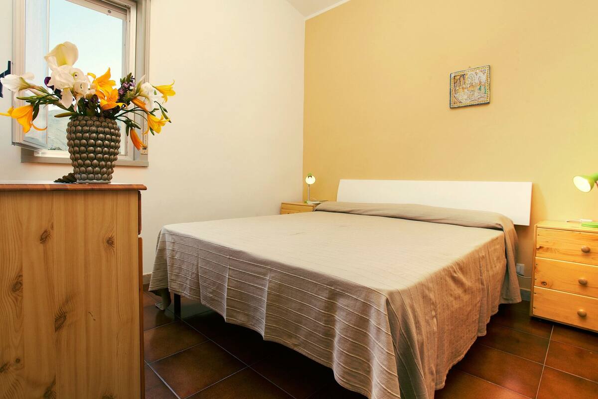 Une chambre à coucher avec un lit, une commode, une table de chevet, une lampe et un vase de fleurs.