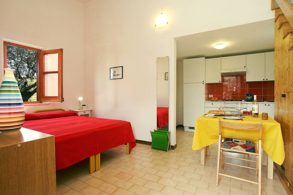 Une pièce de taille modeste comprenant un lit, une petite table recouverte d’une nappe jaune, un coin cuisine et une fenêtre donnant sur des arbres.