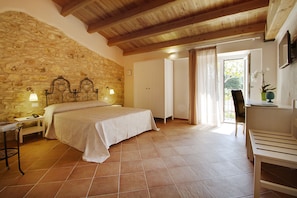 A bedroom with a stone wall, a bed with a patterned bedspread, a bedside table with a lamp, and a chair by the window. - Casale la Macina Hotel & Residence (Castellammare del Golfo)