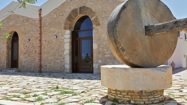 A stone building with arched doorways and a large stone mill in the foreground. - Casale la Macina Hotel & Residence (Castellammare del Golfo)
