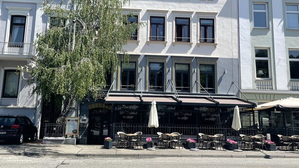 Ein Gebäude mit weißer Fassade, zahlreichen Fenstern und einem roten Ziegeldach. Draußen befinden sich Sitzbereiche mit Sonnenschirmen und Stühlen.