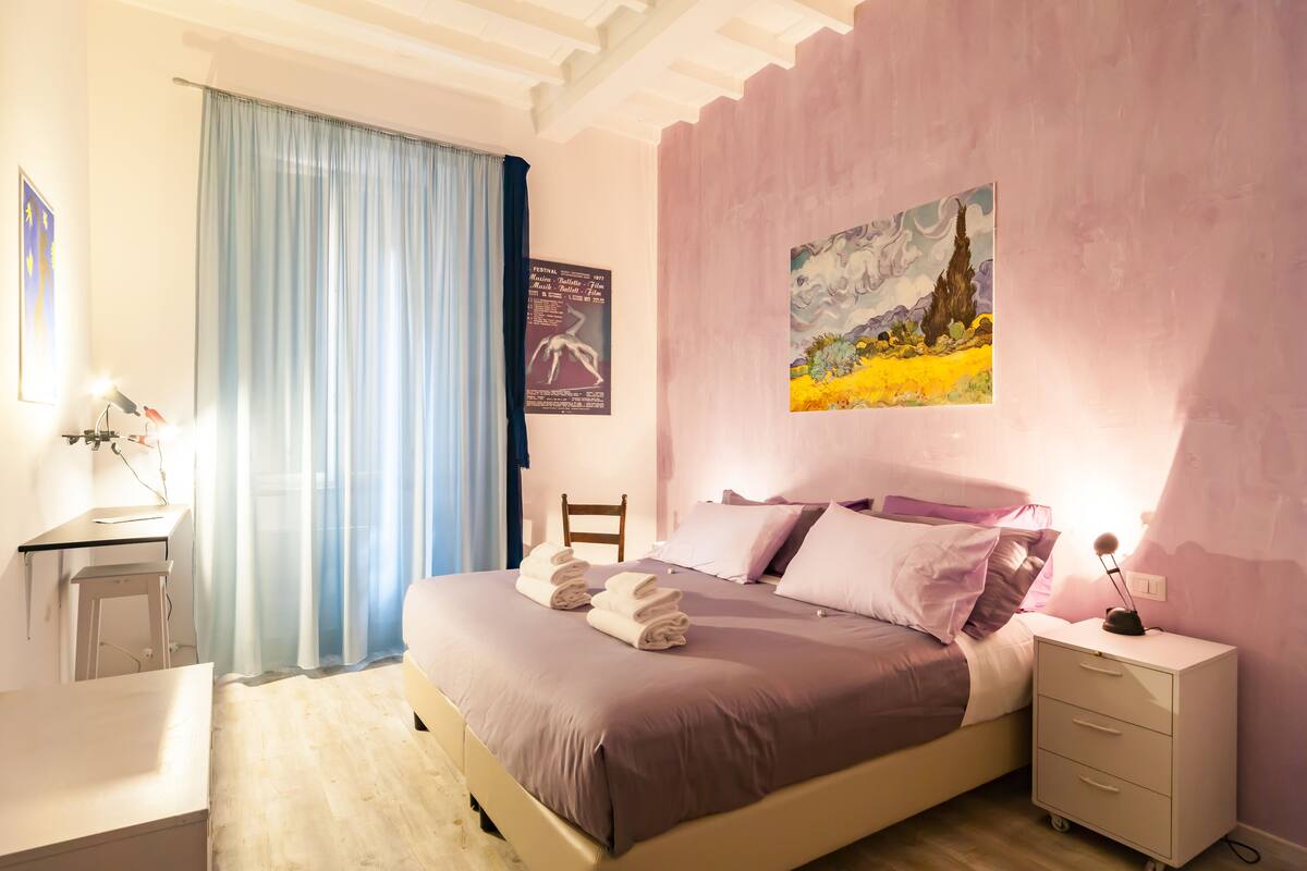 A neatly made bed with pillows and a folded towel, a bedside table with a lamp, a chair, and a wall with a painting.