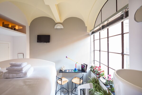 Une chambre d’hôtel comprenant un lit, un bureau avec deux chaises, une télévision et une grande fenêtre agrémentée de plantes.