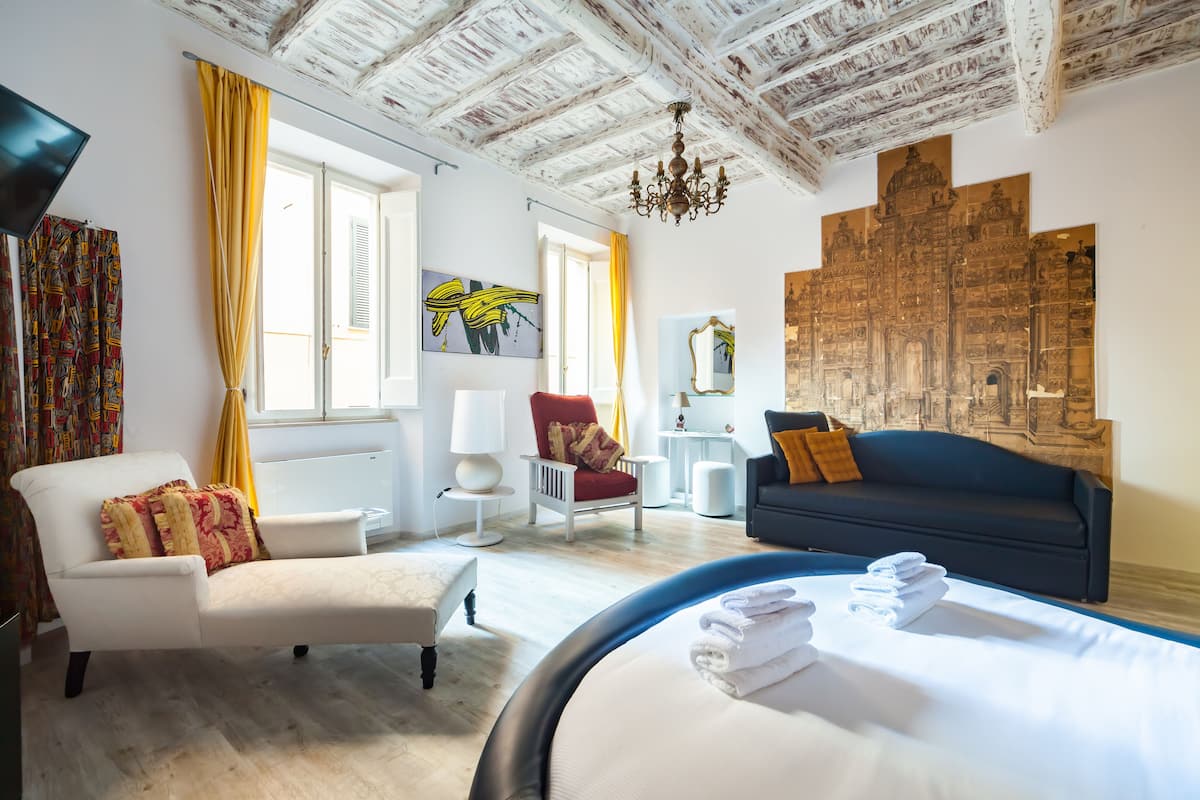 A hotel room with a bed, a sofa, a chair, a desk, and artwork on the walls.