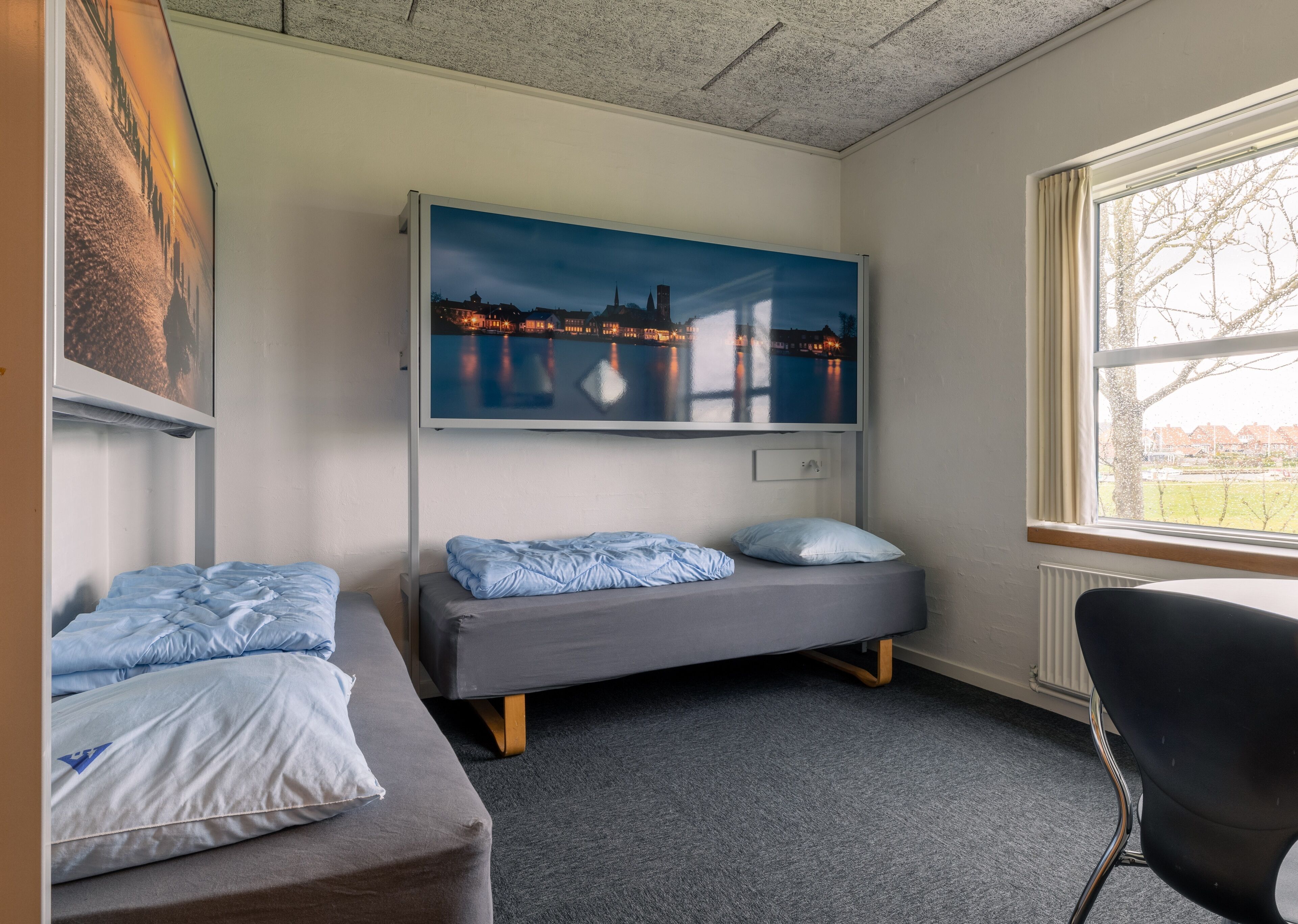 Una habitación con una cama, una silla y un televisor grande en la pared que muestra un paisaje urbano.