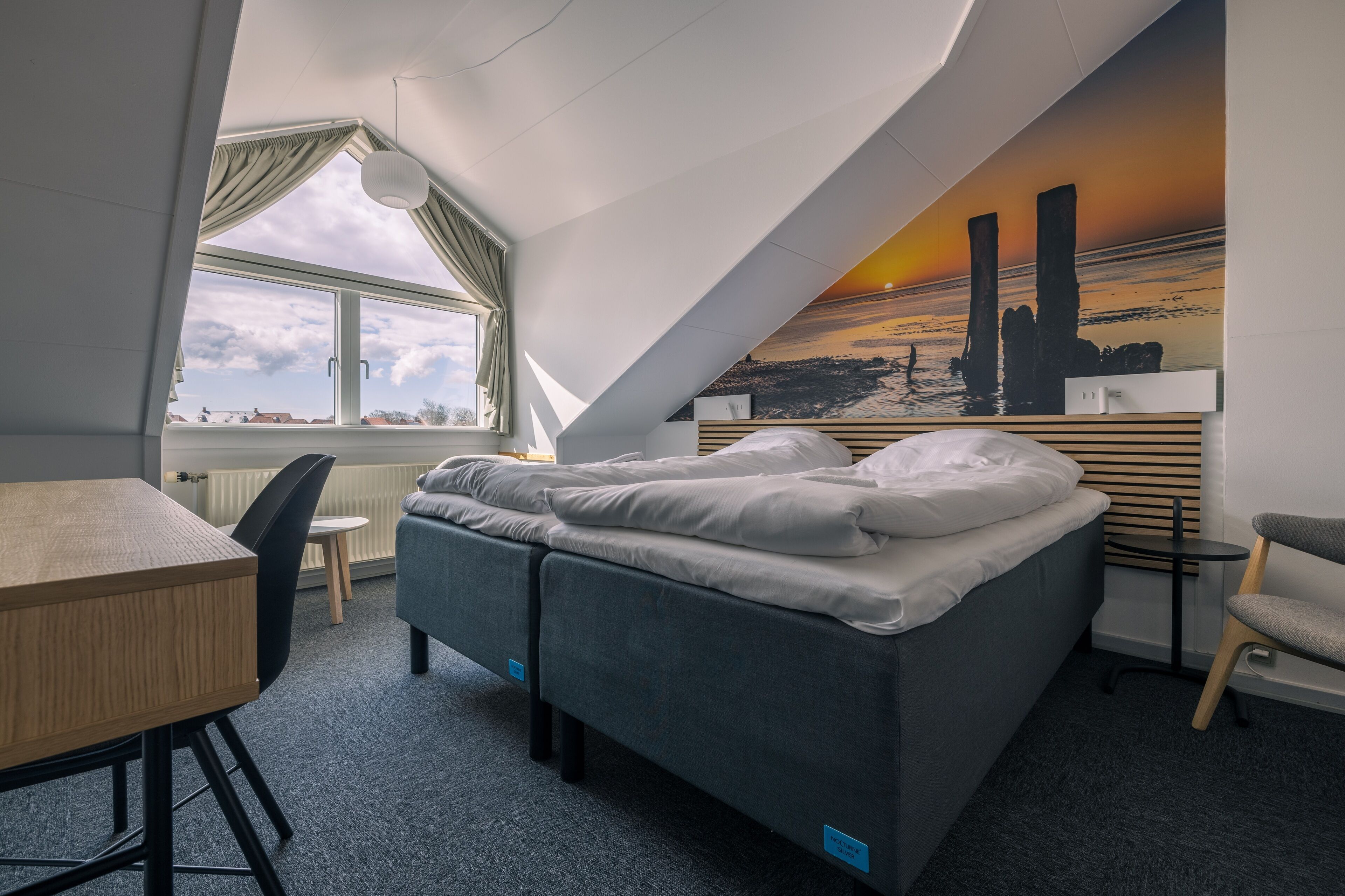 A compact hotel room with a bed, a desk, a chair, and a view of the sunset through the window.