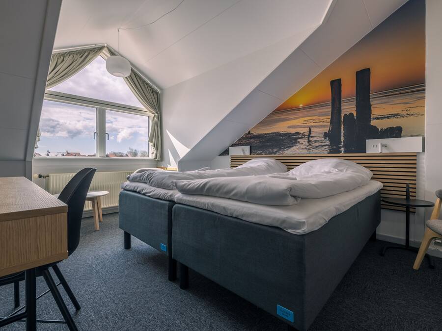 A compact hotel room with a bed, a desk, a chair, and a view of the sunset through the window.