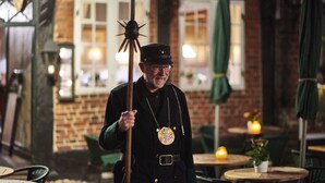 A person in uniform holding a staff in an outdoor setting with brick walls and tables.