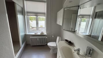 Ein Badezimmer mit weißem Waschbecken, WC und Handtuchheizkörper. Ein Fenster bietet Blick auf grüne Vegetation.