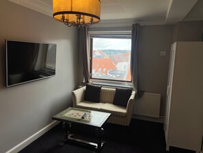A hotel room with a television, a small table, a sofa, a window with a view of buildings, and a radiator. - Det Gamle Rådhus (Grasten)