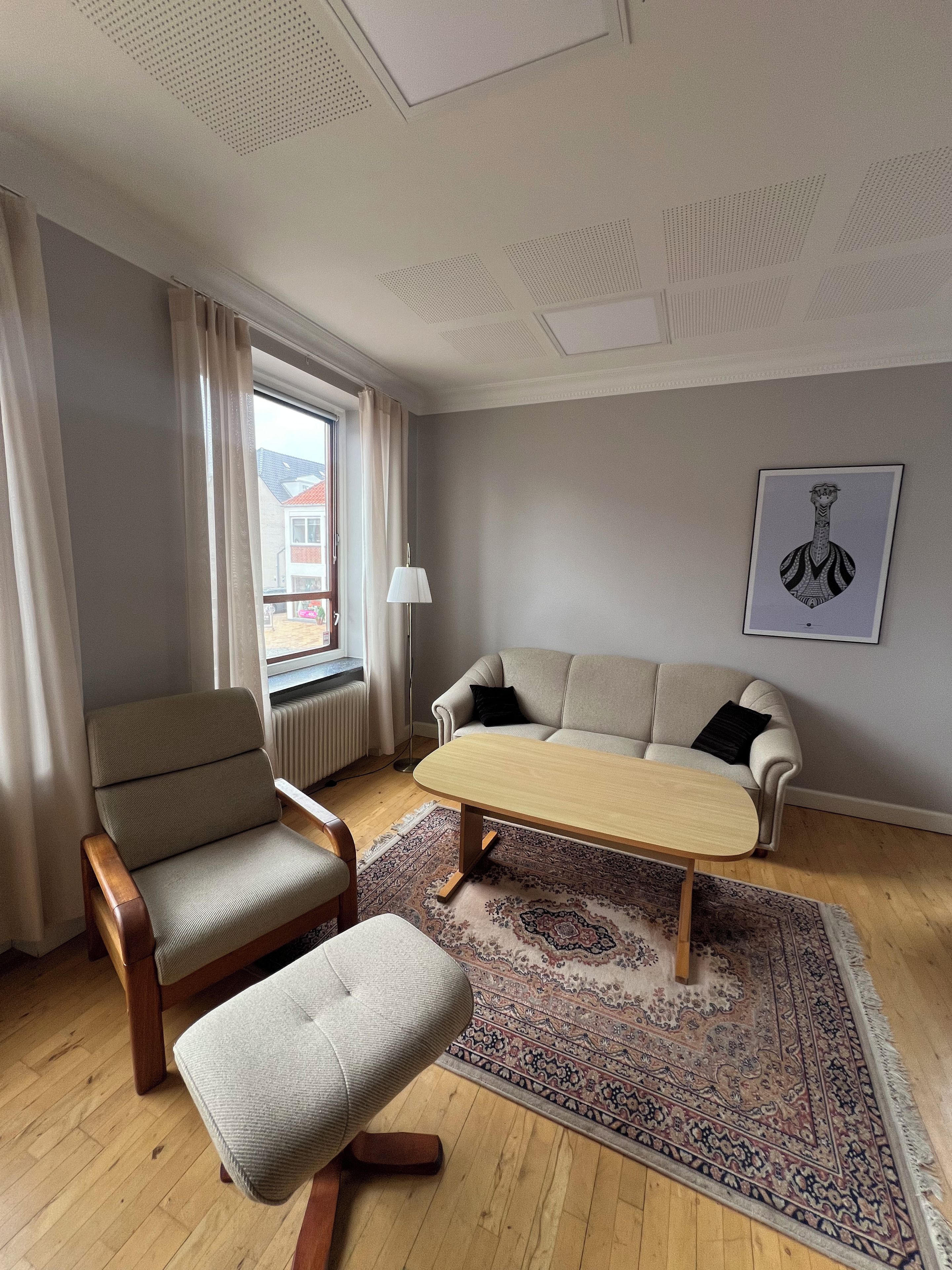 A living room with a sofa, armchair, and ottoman, a patterned rug, a radiator, and a window with curtains.