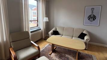 A living room with a sofa, armchair, and ottoman, a patterned rug, a radiator, and a window with curtains.