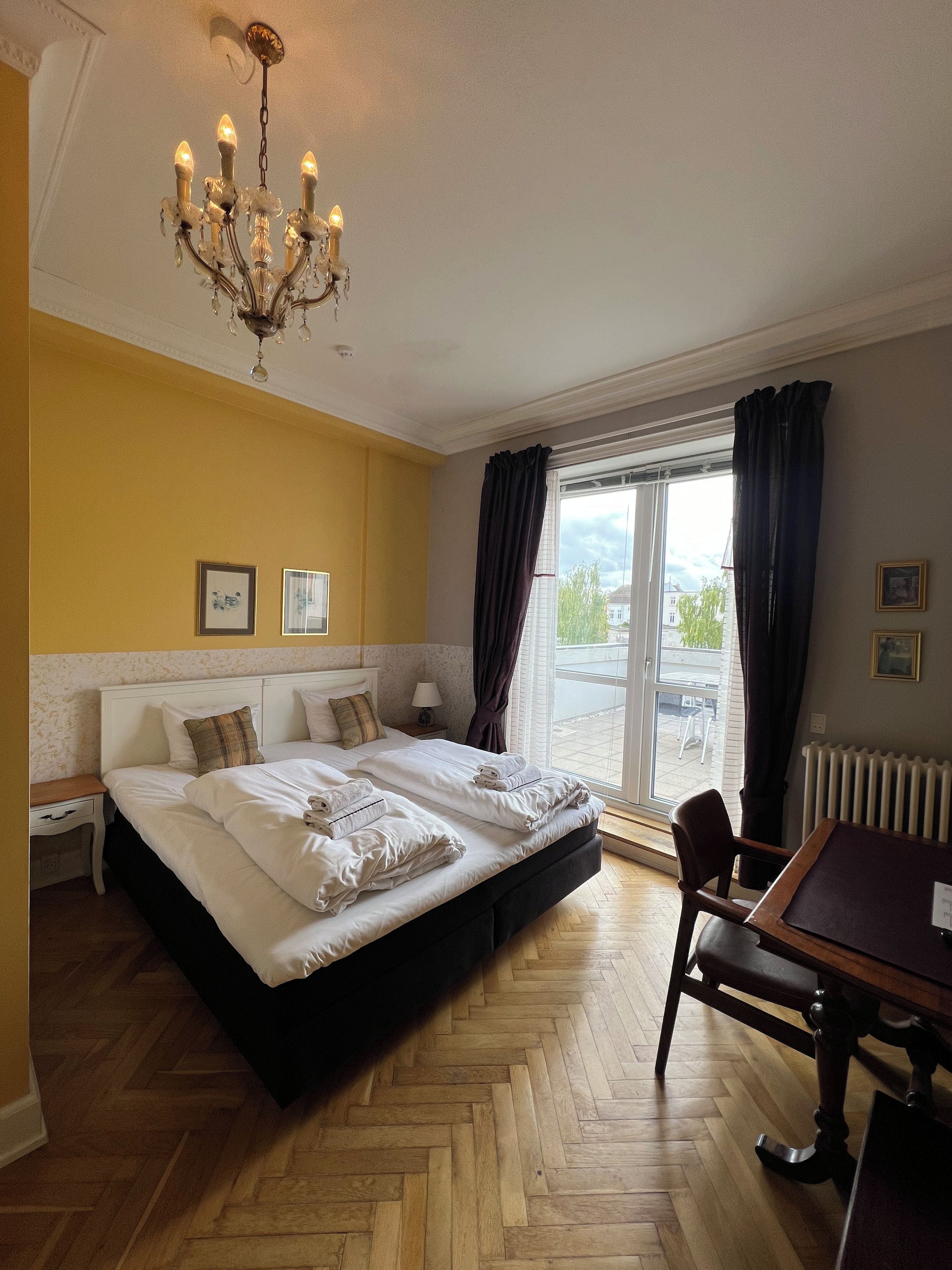 A hotel room with a large bed, a dining table, a chandelier, and a view of the outdoors.