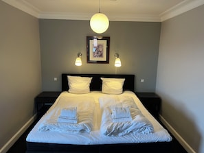 Un lit bien fait, avec du linge de lit et des serviettes blanches, deux tables de chevet, un tableau encadré au mur et un luminaire suspendu.