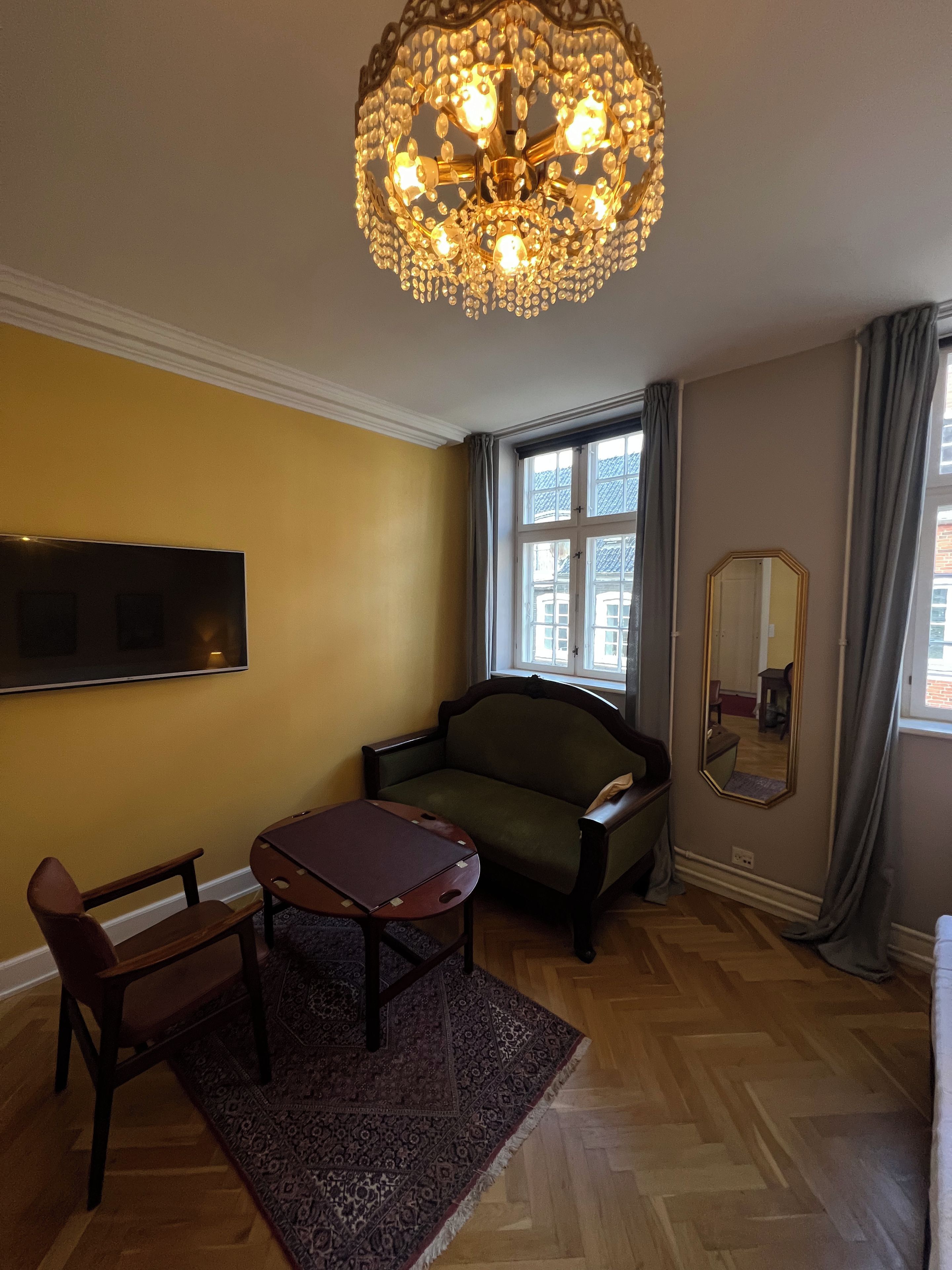 Ett rum med en kristallkrona, en soffa, en fåtölj, ett litet bord, en matta, en tv, ett fönster med gardiner och en spegel.
