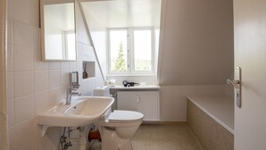 Un baño moderno con un lavamanos, inodoro y bañera blancos. Una ventana proporciona luz natural y vistas a la vegetación exterior.