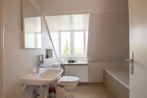 A modern bathroom with a white sink, toilet, and bathtub. There is a window providing natural light and a view of greenery outside. - Det Gamle Rådhus (Grasten)