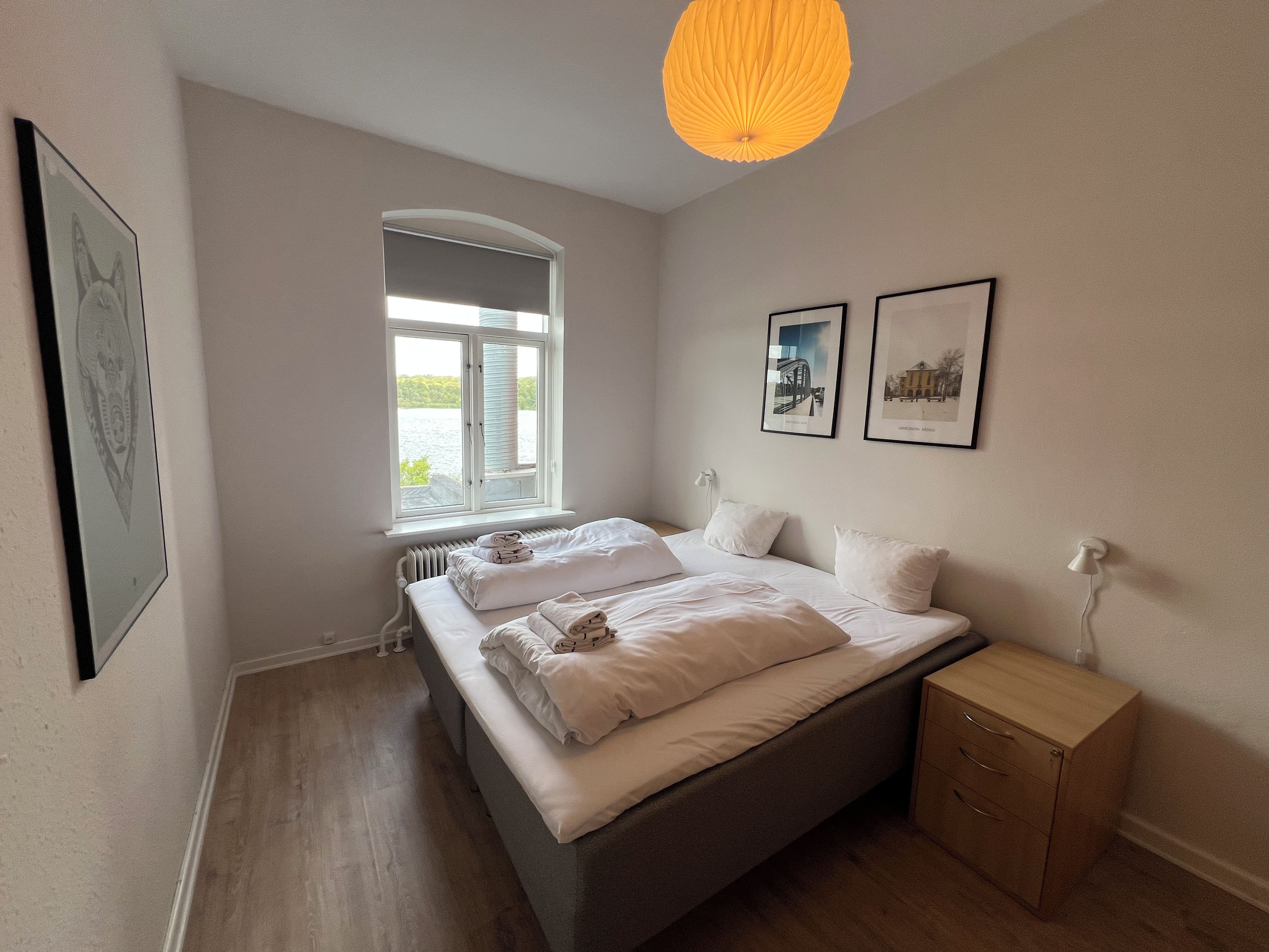A bedroom with a bed, a bedside table, a radiator, a window, and two framed pictures on the wall.
