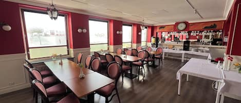 Un comedor con varias mesas y sillas, una zona de bar con estanterías llenas de vasos y botellas, y grandes ventanales que dejan entrar luz natural.