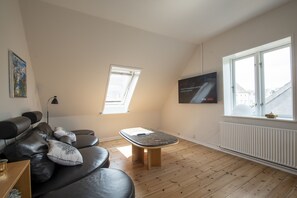 A modern living room with a black leather sofa, a wooden coffee table, a floor lamp, and a large window with a view of buildings. - Det Gamle Rådhus (Grasten)