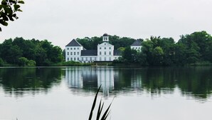 A large, white, multi-story building with a central tower, surrounded by lush greenery and reflecting in calm water. - Det Gamle Rådhus (Grasten)