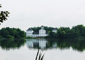 Un grande edificio bianco a più piani, con una torre centrale, circondato da vegetazione lussureggiante e riflesso in acque calme.