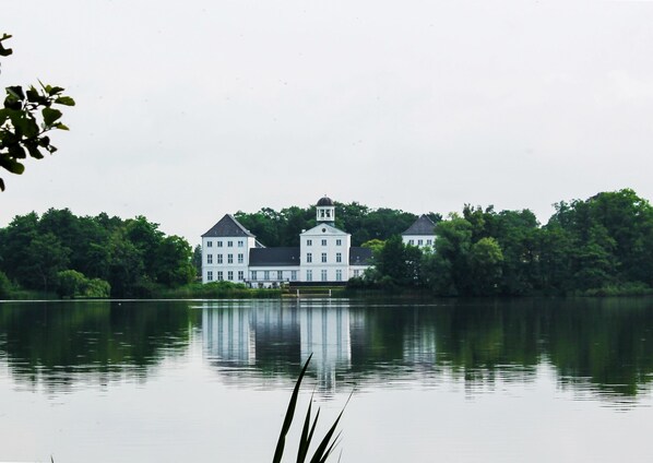 A large, white, multi-story building with a central tower, surrounded by lush greenery and reflecting in calm water. - Det Gamle Rådhus (Grasten)