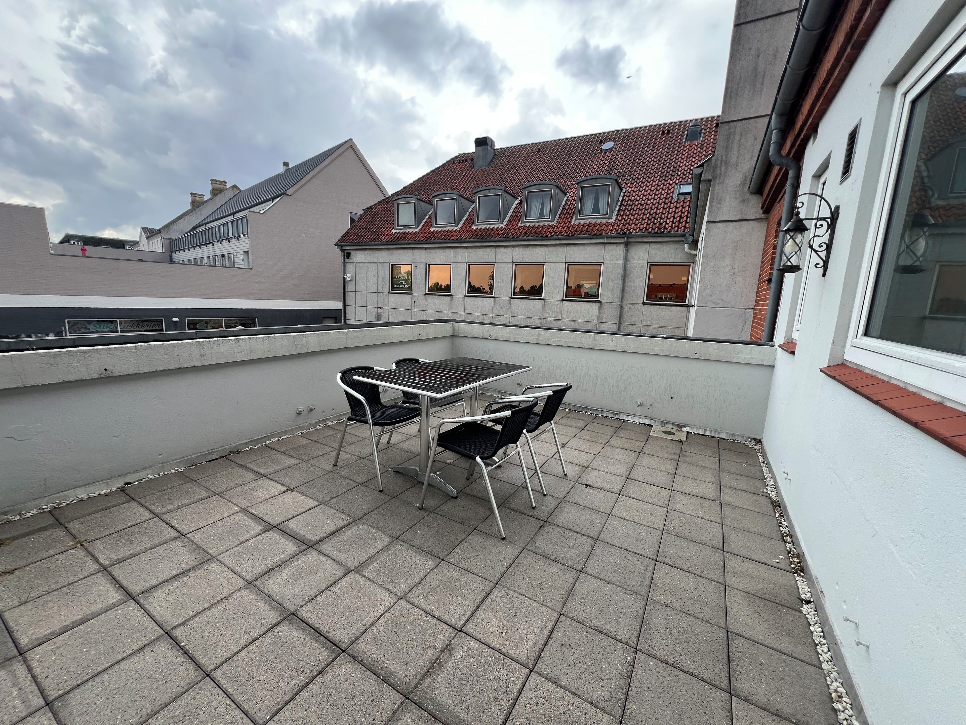 A rooftop terrace with a table and chairs, a view of a building with a red-tiled roof, and a cloudy sky.