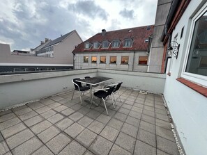 A rooftop terrace with a table and chairs, a view of a building with a red-tiled roof, and a cloudy sky. - Det Gamle Rådhus (Grasten)
