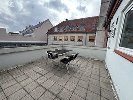 Una terraza en la azotea con mesa y sillas, vista a un edificio con tejado de tejas rojas, y un cielo nublado.