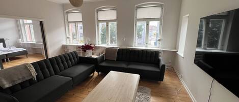 A modern living room with a sectional sofa, a coffee table, and a TV. There are large windows with blinds, a ceiling light, and a radiator.