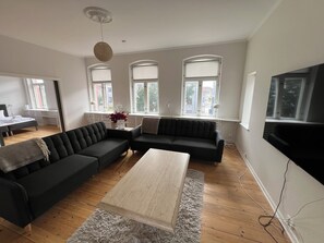 Un salon moderne avec un canapé d’angle, une table basse et un téléviseur. De grandes fenêtres sont équipées de stores, et on y trouve également un luminaire au plafond et un radiateur.