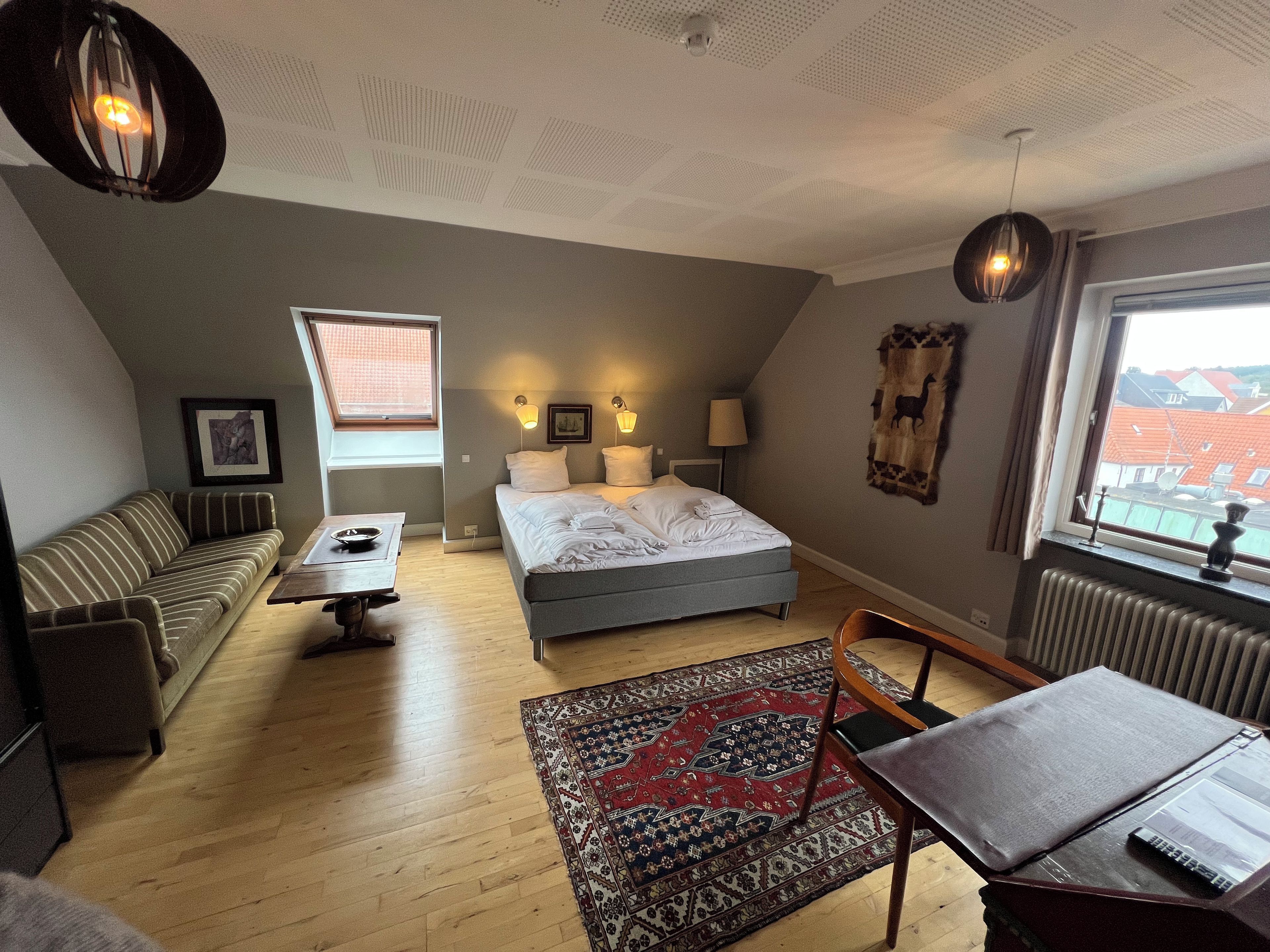 A bedroom with a bed, a sofa, a table, a chair, a rug, and a window with a view of buildings.