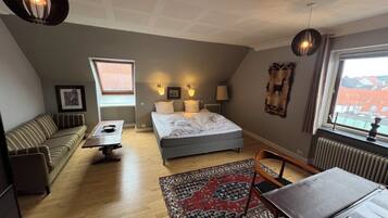 A bedroom with a bed, a sofa, a table, a chair, a rug, and a window with a view of buildings.