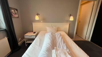 Habitación de hotel con una cama individual, una mesita de noche, una lámpara y una puerta que da a otra habitación.