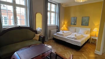 A hotel room with a bed, a sofa, a table, and a chair. There are two windows with curtains, a radiator, and a chandelier.